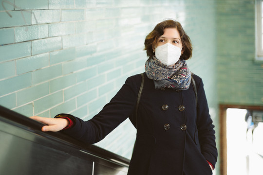 Eine Frau fährt auf einer Rolltreppe nach oben und trägt dabei eine FFP2-Atemschutzmaske. Im Hintergrund eine gekachelte Wand in einer Ubahn-Station. fotografixx via Getty Images
